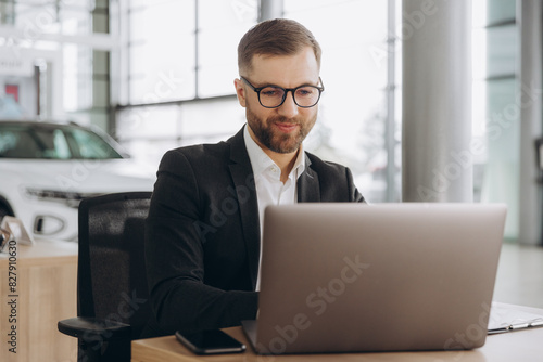 Smiling bearded salesman in suit and glasses typing on his laptop at new cars showroom