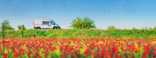 panorama landscape with field of red poppies and white van on road under blue sky with copy space