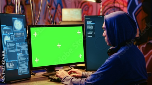 Asian hacker in hidden place with graffiti walls using green screen computer to deploy malware on unsecured devices, stealing sensitive data from unaware users online and selling it on black market photo