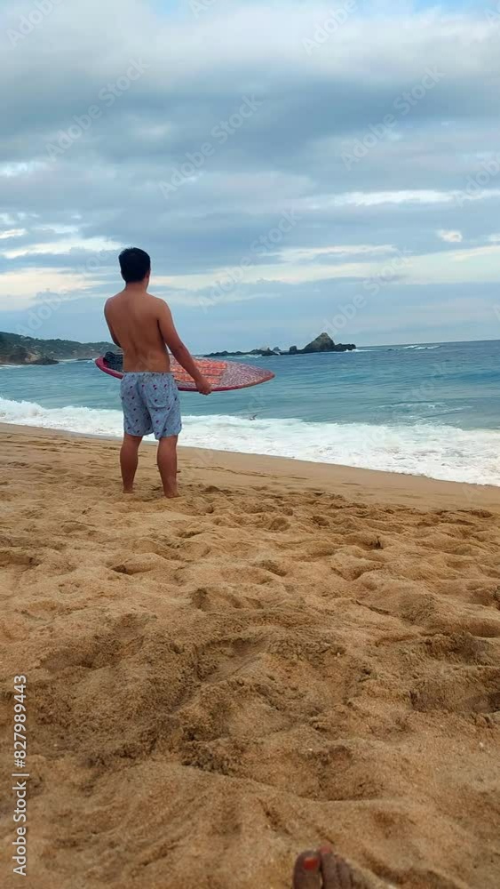 young latin man watching a skimboarder catching a wave on the seashore, vertical, static young latin man watching a skimboarder catching a wave on the seashore, vertical, static