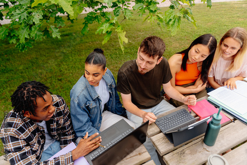 Multiracial group of five students sitting together at a table in the university campus with academic materials, laptops and notes working on a class project. Education and academic year