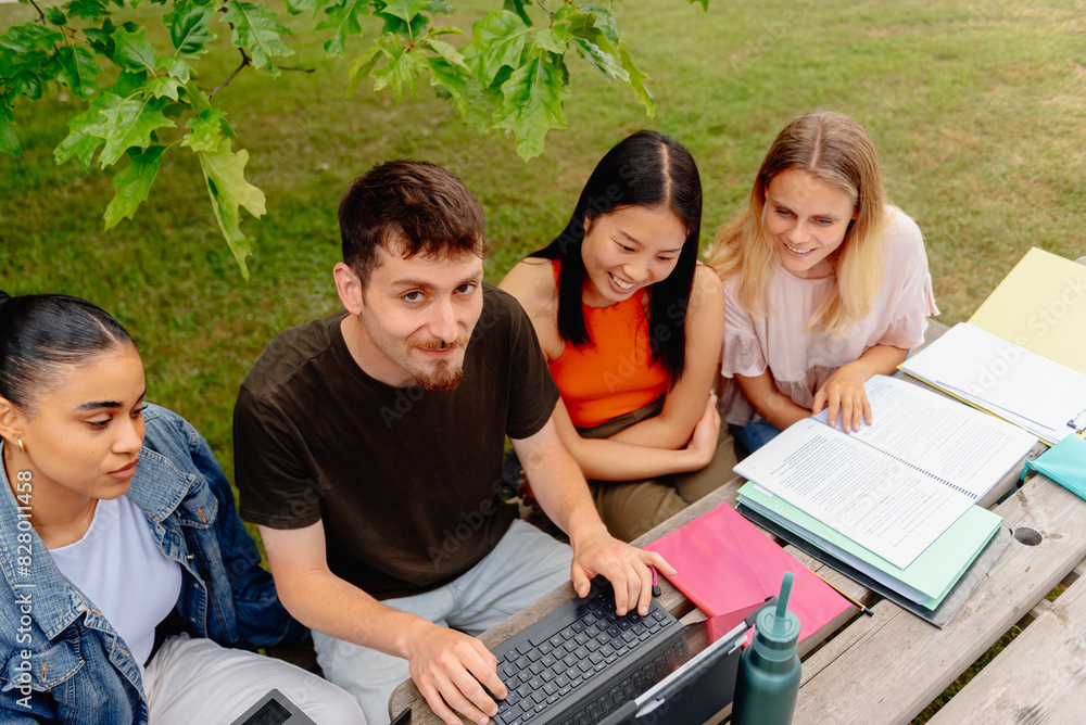 Young Caucasian male using a laptop computer with his multiracial group of students seated at a table outside the university campus. Classmates studying together Young Caucasian male using a laptop computer with his multiracial group of students seated at a table outside the university campus. Classmates studying together
