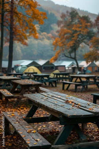 Autumn camping site with wooden picnic tables and colorful fallen leaves, surrounded by vibrant trees and cozy tents in the background