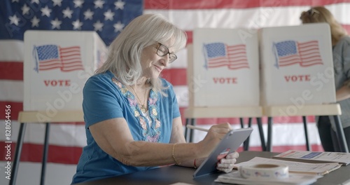 Medium shot, elderly woman election worker writes on tablet computer at polling station. Young woman votes at booth in background with US flag behind
