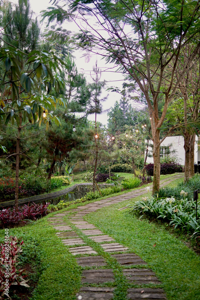 A stone path winds through a lush garden, lit by string lights. It leads towards a hidden pond, with a white building nestled among the trees.