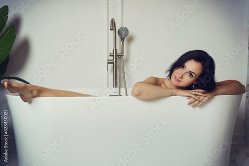 charming brunette woman in a black dress is sitting in the bathtub with her leg out photo