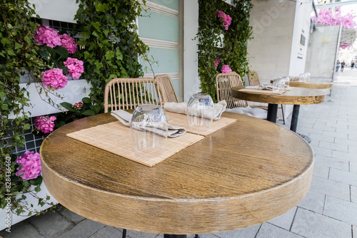 Empty wooden tables and chairs with pink decorative flowers adorn the facade of a summer street cafe