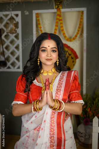 Portrait of beautiful young Indian woman elegantly attired in saree, adorned with gold jewellery and bangles, standing with her hands in a namaste gesture.