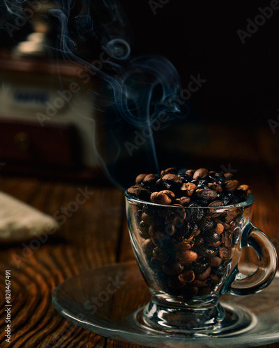 Coffee beans of varying degrees of roasting in a transparent coffee cup on a wooden table with manual coffe grinder and smoke in the background