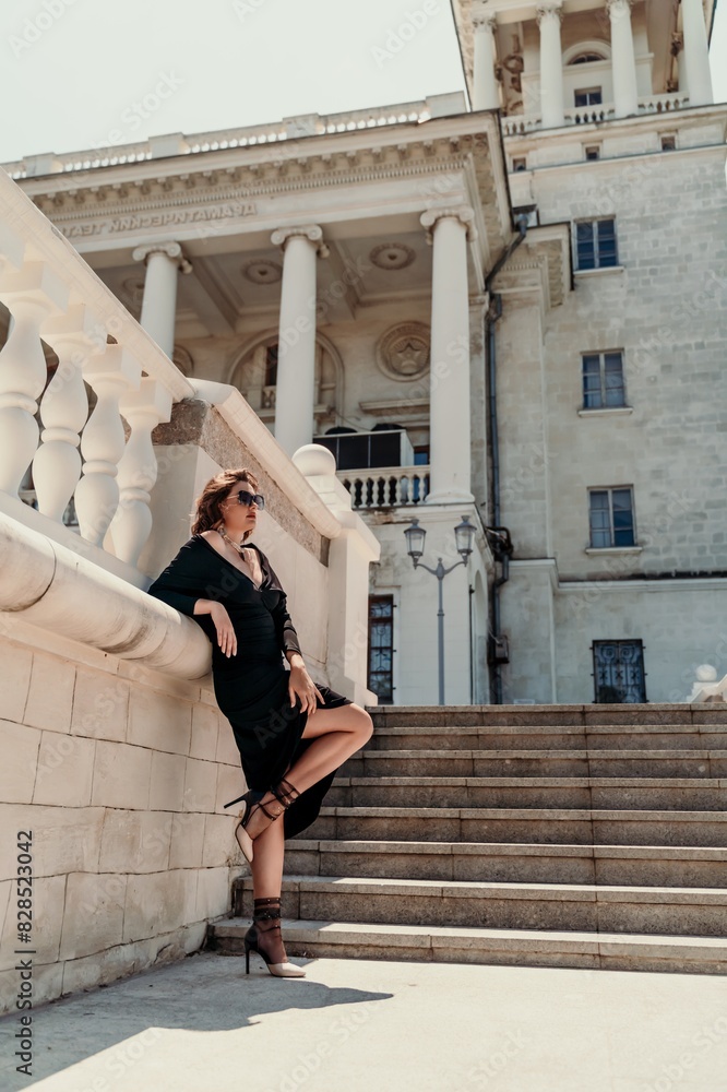 Stylish woman in the city. Fashion photo of a beautiful model in an elegant black dress posing against the backdrop of a building on a city street