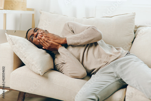 Tired African American man suffering from a severe headache while sitting on a couch at home He appears stressed, sick, and frustrated, reflecting the pain and discomfort caused by the migraine The