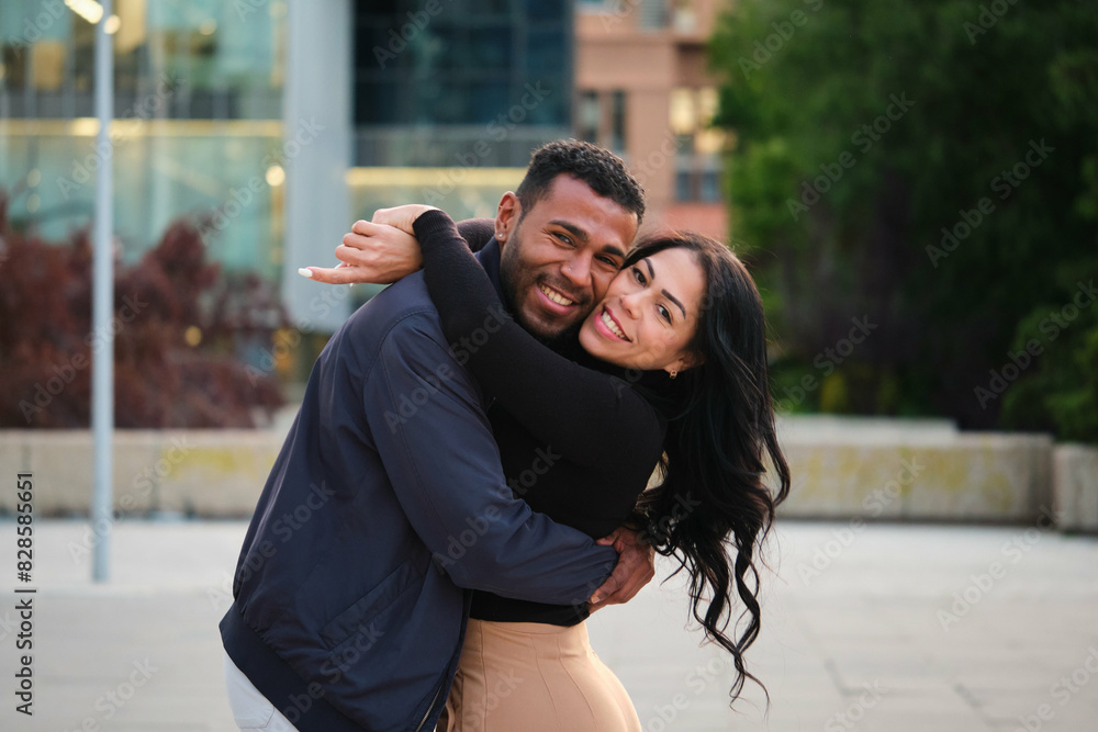 Latin man and woman are hugging each other in a city. They both have smiles on their faces, indicating that they are happy