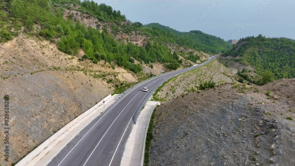 Aerial view of a white car driving on a winding highway through rocky mountainous terrain with green vegetation Aerial view of a white car driving on a winding highway through rocky mountainous terrain with green vegetation