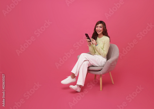 Full length beautiful Asian teen woman using smartphone and sitting on chair isolated on pink background. People lifestyle Technology Concept.