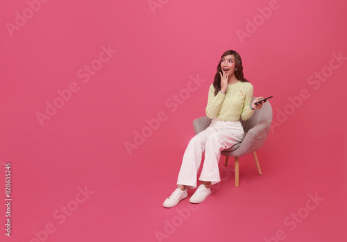 Full length excited beautiful Asian teen woman using smartphone and sitting on chair isolated on pink copy space background. People lifestyle Technology Concept.