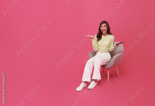 Full length beautiful Asian teen woman using smartphone and sitting on chair isolated on pink copy space background. People lifestyle Technology Concept..