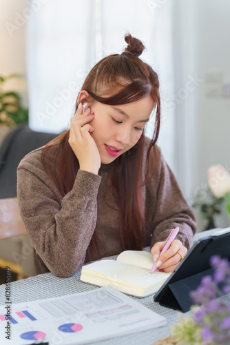 Asian business woman in sweater watching entertainment video on tablet and thinking about business plan to taking notes during planning about marketing strategy of new startup while working from home photo