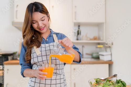 Young asian woman pouring orange juice from jug into the glass while preparing vegetables salad and cooking healthy breakfast food for homemade snack in modern kitchen with healthy lifestyle at home