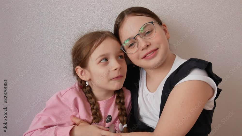 Two little happy sisters hugging cuddled up to each other sitting on the bed. The love of brothers and sisters. A schoolgirl in glasses and a school uniform. A preschooler in a pink sweater. Two little happy sisters hugging cuddled up to each other sitting on the bed. The love of brothers and sisters. A schoolgirl in glasses and a school uniform. A preschooler in a pink sweater.