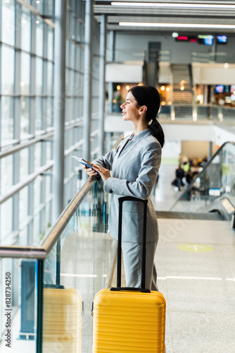 Asian girl in business suit at airport terminal with yellow suitcase looking in window. Business woman with valise looking in panorama window while waiting on flight boarding for vacation.