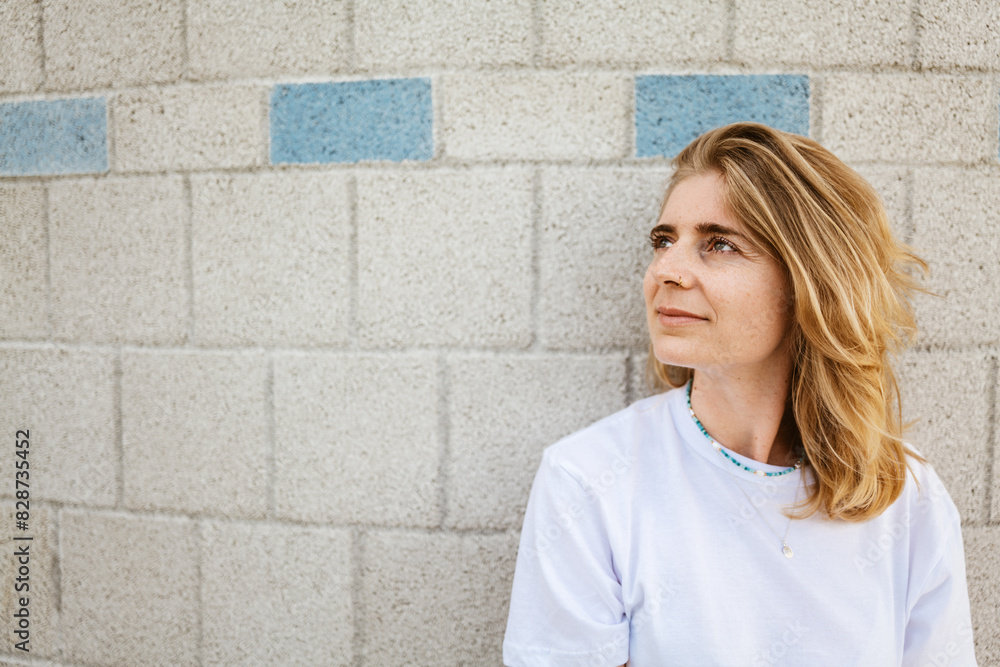 Blond-haired woman in casual white T-shirt looking thoughtful against a brick wall with blue accents. Blond-haired woman in casual white T-shirt looking thoughtful against a brick wall with blue accents.