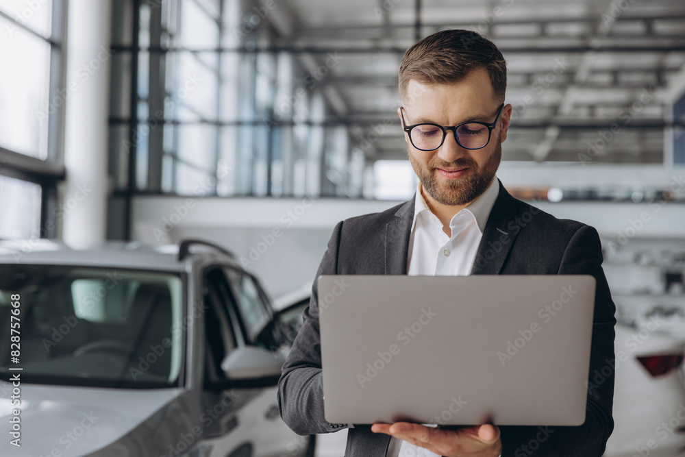 Modern bearded man in glasses and suit vehicle sales consultant using laptop inside car dealership Modern bearded man in glasses and suit vehicle sales consultant using laptop inside car dealership
