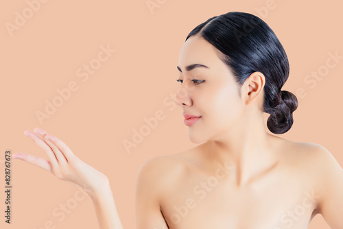A young woman with smooth, glowing skin poses against a peach-colored background. She is looking at her open hand with a gentle smile. Her hair is neatly tied back, emphasizing her natural beauty