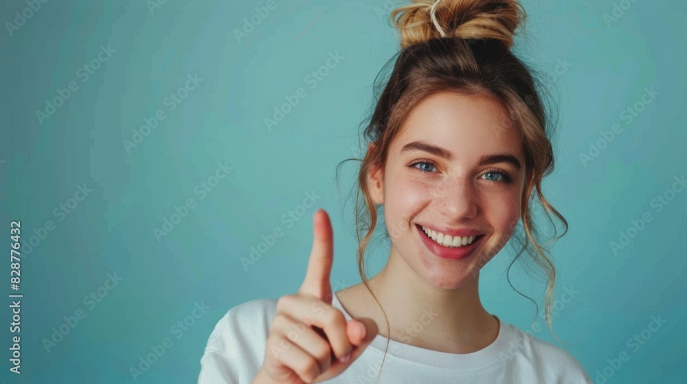 A smiling young woman with a messy bun hairstyle points upward with her index finger against a teal background. A smiling young woman with a messy bun hairstyle points upward with her index finger against a teal background.