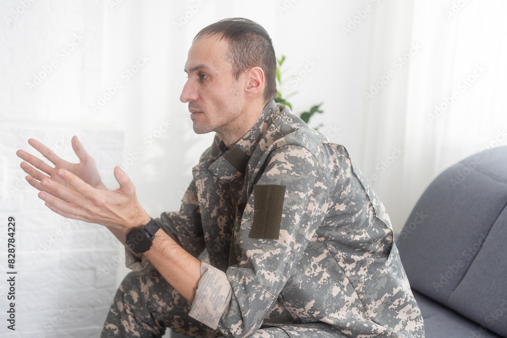 Fototapeta premium Portrait of calm serious Caucasian military man wearing camouflage uniform and cap sitting on a sofa, having depressed facial expression.