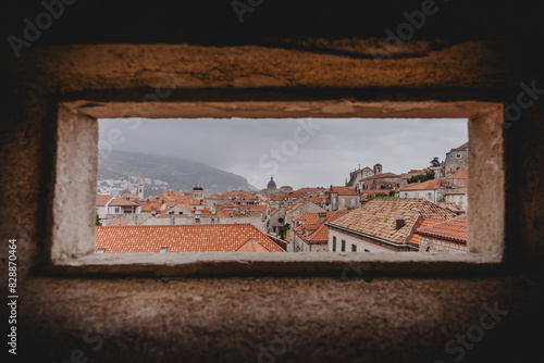 Historic view of Dubrovnik’s red rooftops and old-world architecture framed through an ancient stone window. The misty backdrop and timeless charm of this Croatian city are captured beautifully.