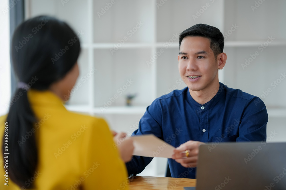 A man in a blue shirt handing a document to a woman in a yellow jacket during a meeting in a modern office setting with a laptop on the desk. A man in a blue shirt handing a document to a woman in a yellow jacket during a meeting in a modern office setting with a laptop on the desk.