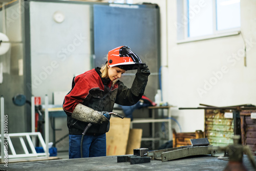Beautiful blonde woman works as a welder in workshop, operating welding machine, wearing protective clothing and a welding mask.