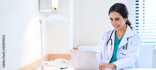 Fotografie Smiling professional female doctor in white medical coat working with laptop computer, sitting at the table near the window in hospital office, looking at laptop screen