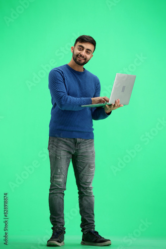 A man, full-length, on a green background, uses a laptop © Katsiaryna