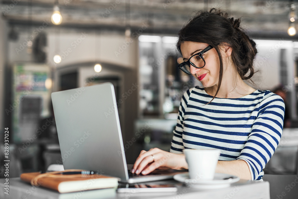 Woman working remotely from coffee shop. Cappucino and laptop on table. Female freelancer working on notebook in cafe. Woman working remotely from coffee shop. Cappucino and laptop on table. Female freelancer working on notebook in cafe.