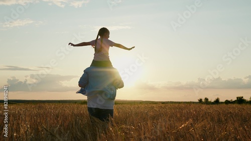 Little daughter on shoulders of her father. Girl and dad are traveling through wheat field. Child and parent play in nature. Happy family, childhood concept. Slow motion. Daughter, dad farmer walking photo