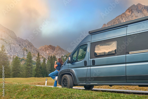 Woman leaning against a camper van, enjoying a scenic mountain view with rocky peaks and pine trees in the background. The scene captures the essence of adventure, freedom, and outdoor travel.