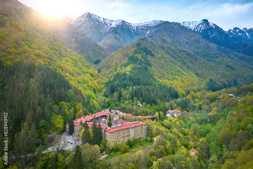Aerial view of Rila Monastery nestled in the lush green forests of Rila Mountain with snow-capped peaks in the background  Bulgaria. The historical complex features red-roofed buildings.
