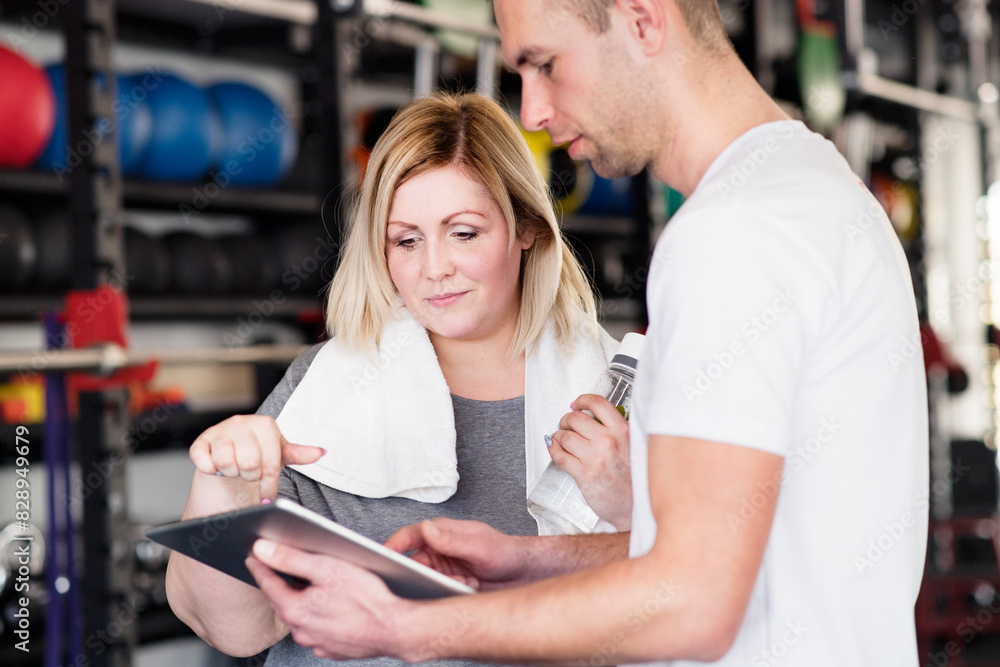 Overweight woman consulting with trainer, discussing daily workout plan. Personal trainer talking with client, discussing exercise program in gym.