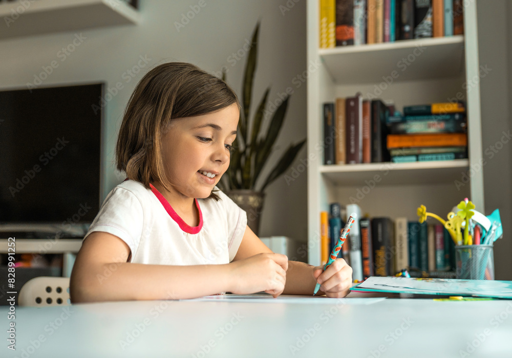 Little girl wries something with left hand at a desk in front of a book shelves. Children education concept. Little girl wries something with left hand at a desk in front of a book shelves. Children education concept.
