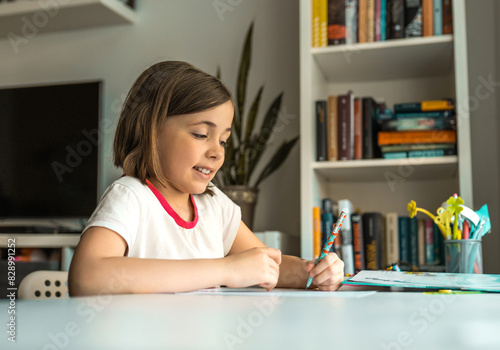 Little girl wries something with left hand at a desk in front of a book shelves. Children education concept.