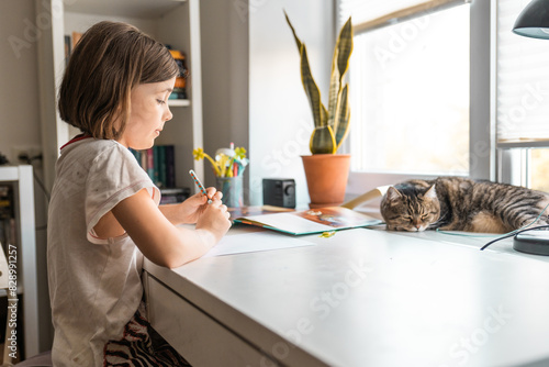 Little girl wries something with left hand at a desk at home with a cat. Children education concept.