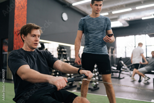 Fotografie Focused beginner sportsman with personal instructor working out in gym by using rowing machine