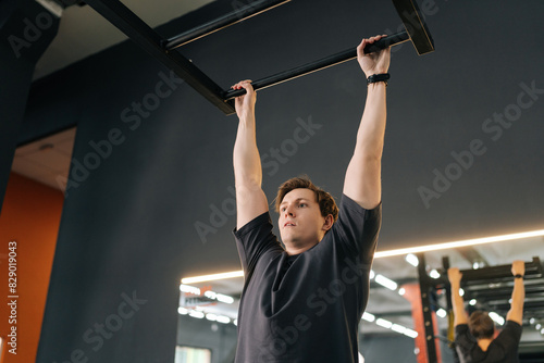 Exhausted muscular male hanging on bar and does one pull-up during sport workout training in modern gym. Tired motivated beginner sportsman showing hard expression on face in pulling up own body.