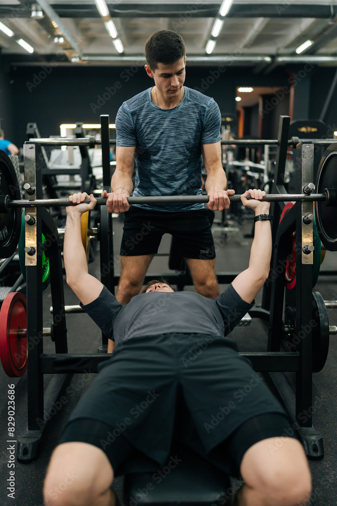 Vertical shot of experienced fitness instructor helping beginner sportsman doing barbell bench press exercise during personal workout in gym. Male doing barbell bench press under couch supervision Vertical shot of experienced fitness instructor helping beginner sportsman doing barbell bench press exercise during personal workout in gym. Male doing barbell bench press under couch supervision