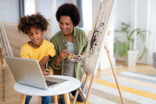 Smiling African American mother and daughter painting together and using laptop in a living room. photo
