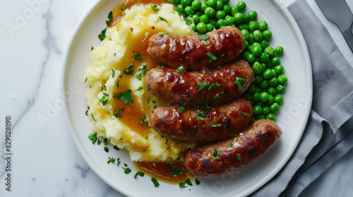 Pork sausages with creamy mashed potatoes, gravy sauce, green peas and greenery on white plate on dark grey background and napkin overhand photo