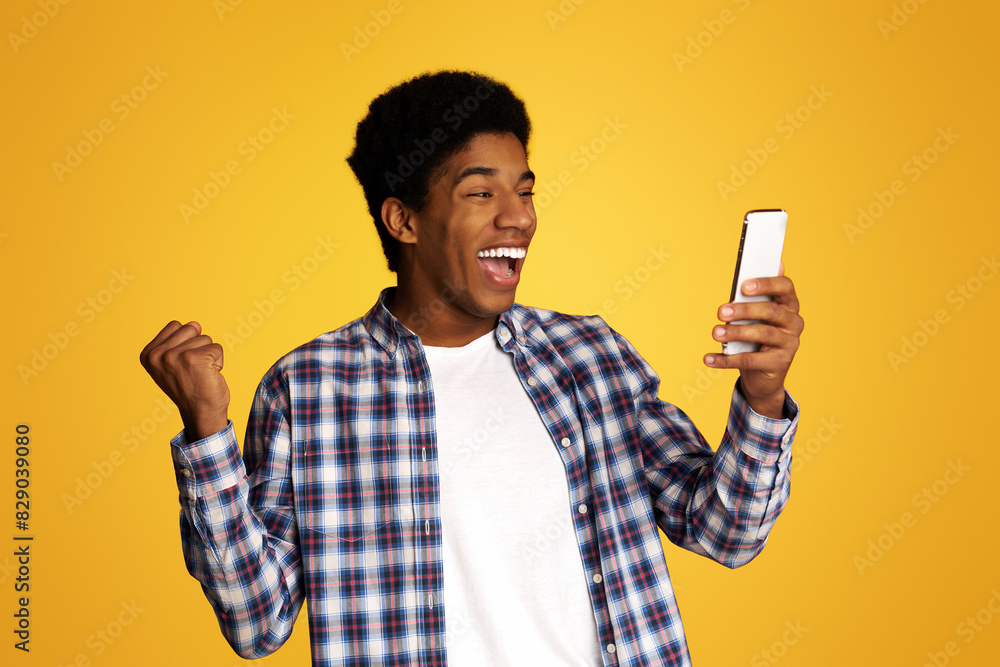 Winner Concept. African Guy Using Smartphone over Yellow Studio Background Winner Concept. African Guy Using Smartphone over Yellow Studio Background