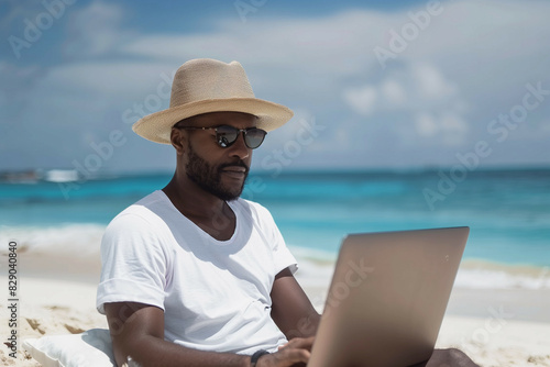 A man sitting on a beach wearing a straw hat and sunglasses, working on a laptop with a serene ocean view in the background
