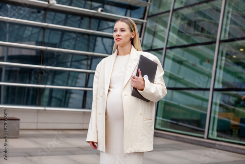 Beautiful pregnant blonde businesswoman in a white business suit with a blazer stands outdoors with a laptop near a corporate building, showcasing professional and modern maternity style